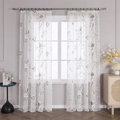 A pair of sheer curtains with a floral pattern hanging in front of a window, with a small table and a cabinet visible in the room.