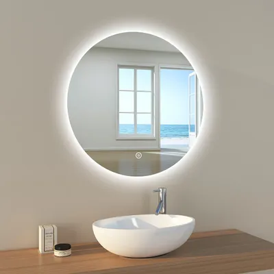 A round LED bathroom mirror with a light ring is mounted on a wall above a white vessel sink. The mirror reflects a room with large windows opening to a sunny ocean view. A faucet is visible above the sink, and a small box and jar are on the wooden counter