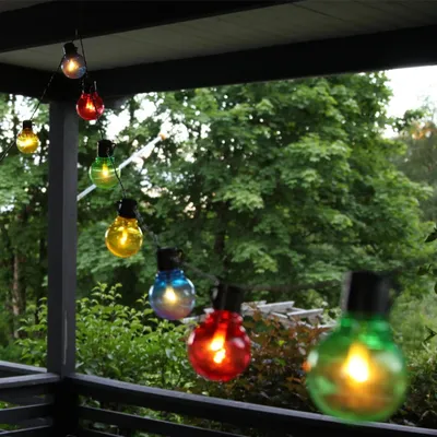 A string of colorful LED party lights hangs on a porch or balcony, with lush green trees in the background.