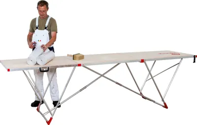 A man in overalls is standing next to a long, foldable table, holding two rolls of white material.