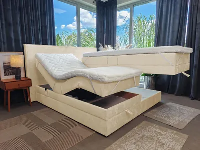 A beige adjustable bed with storage is shown in a room with large windows and dark curtains.