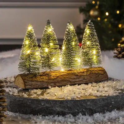 A decorative Christmas display featuring four miniature trees on a log base, illuminated by warm white LED lights, set against a blurred background with bokeh lights.