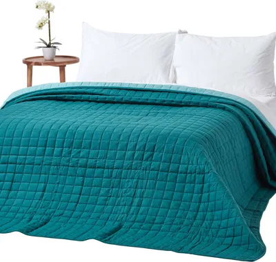 A teal quilted bedspread is draped over a bed with white pillows. A small wooden side table with a potted orchid is visible to the left.