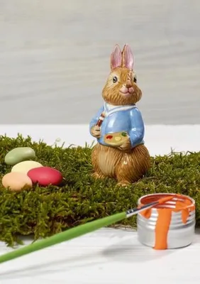 A ceramic bunny figurine dressed in a blue jacket, holding a palette and paintbrush, surrounded by moss and Easter eggs, with a can of orange paint and a paintbrush in the foreground.