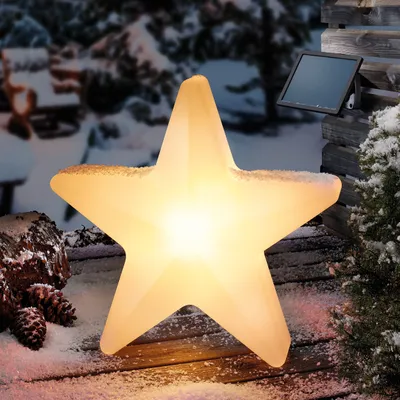 A glowing star-shaped light sits on a wooden surface covered in snow, with pine cones and a snowy tree in the background. A solar panel is attached to a wooden structure behind the star.