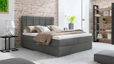A bedroom scene featuring a large grey box spring bed with pillows and a blanket, set against a brick wall.