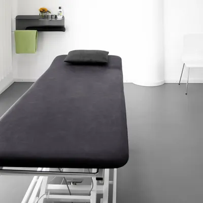 A treatment table with a dark grey cover and a dark grey pillow, placed in a clean, modern room with a shelf holding a green towel, a flower, and bottles.