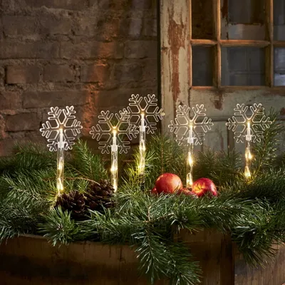 Four snowflake-shaped LED lights are placed among pine branches, with two red apples and pinecones.