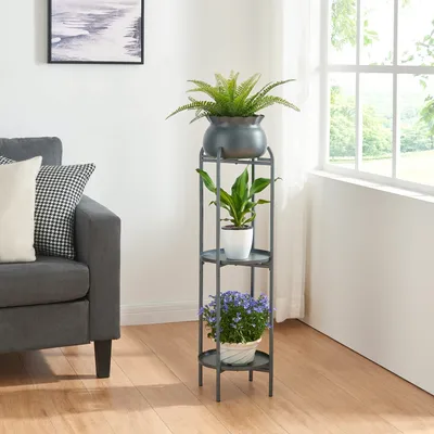 A plant stand with three plants is placed next to a sofa in a living room with a window in the background.