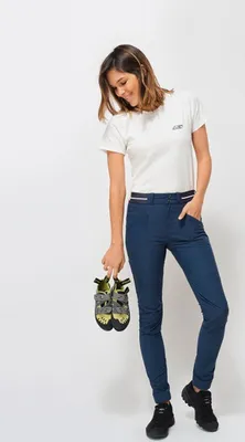 Dame trägt die "Pro Model Hose" von "Looking for Wild" in Blau; hält Kletterschuhe in der Hand; lächelt.