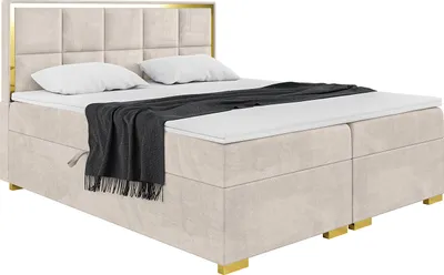 A product shot of a beige upholstered boxspring bed with a gold frame and headboard, set against a white background.