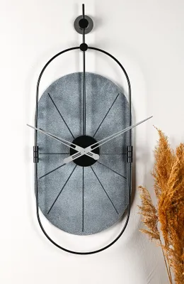 A modern, oval-shaped wall clock with a metallic finish and black metal frame, hanging on a white wall next to a dried plant.
