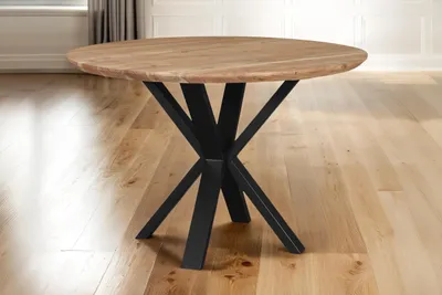 A round wooden dining table with a black spider leg base, set on a wooden floor.