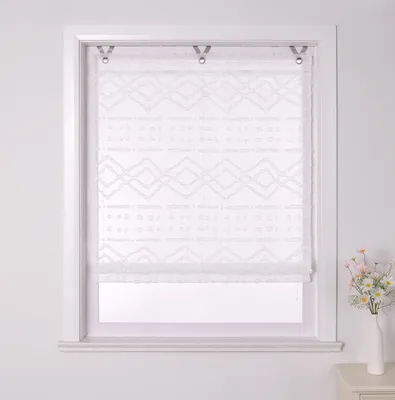 A white sheer Roman shade with a geometric pattern is installed in a window frame. A vase with flowers is on the right side of the image.