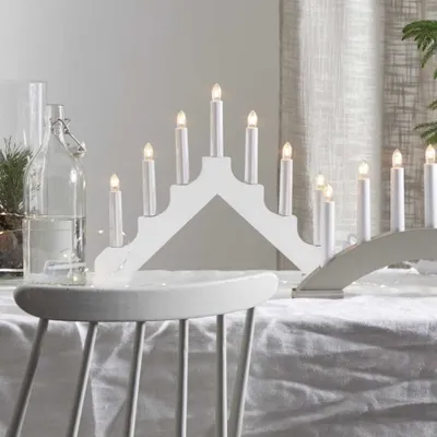 A white candle bridge light is displayed on a table covered with a white tablecloth, with a glass bottle and greenery in the background.