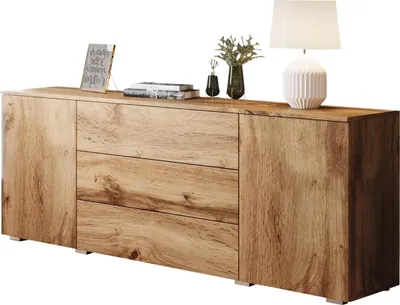 A wooden sideboard with three drawers and two doors, displayed against a white background. Decorative items including a framed picture, books, a vase with greenery, and a lamp are placed on top.