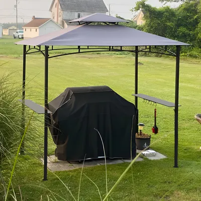 A BBQ grill covered by a black tarp is placed under a gray gazebo in a grassy yard.