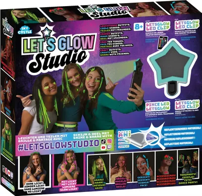 The image shows the packaging of a "Let's Glow Studio" craft and styling set, featuring three young girls with glowing hair and face paint, posing for a selfie with an LED clip attached to a phone. The packaging highlights the product's features and includ