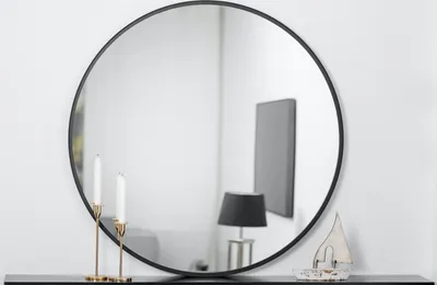 A round black-framed mirror is displayed on a white wall, reflecting a lamp and a framed picture. Two white candles in gold holders are in the foreground, and a small decorative boat is on the right.