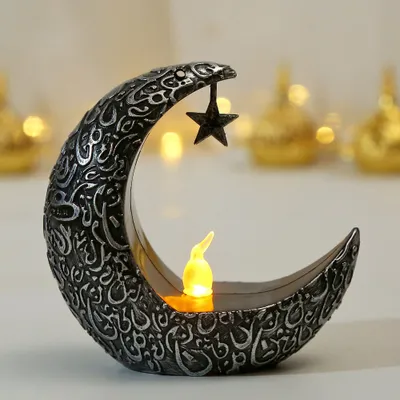 A decorative crescent moon with a star hanging from it, featuring an LED candle inside. The background is blurred with warm, golden lights.