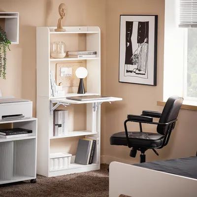 A home office setup with a white folding desk and shelves, a black office chair, and artwork on the wall.
