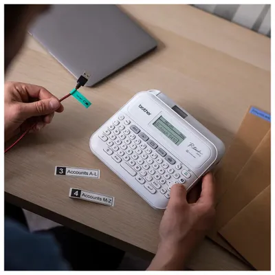 A person is using a Brother P-touch D410 label maker to label a red cable connected to a laptop.