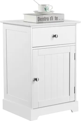 A white nightstand with a drawer and a cabinet door, topped with a book and a teacup.
