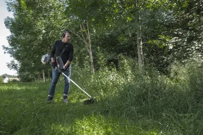 A man is using a string trimmer in a grassy area with trees in the background.
