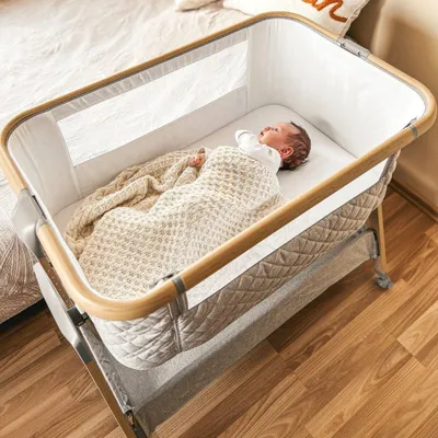A newborn baby sleeping in a bedside bassinet next to a bed.
