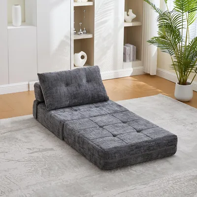 A gray floor sofa with a pillow is placed on a rug in a room with a bookshelf in the background.