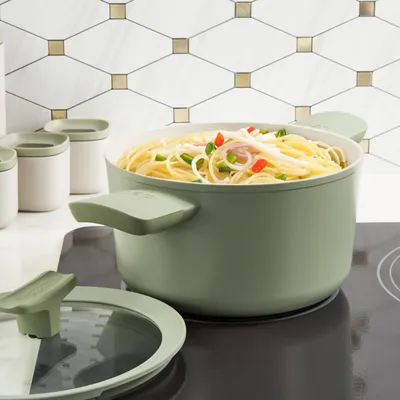 A sage green pot filled with spaghetti and vegetables sits on a black stovetop, with a glass lid and other kitchen containers in the background.
