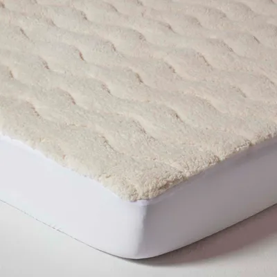 A close-up, angled view of a cream-colored fleece mattress topper with a textured, wavy pattern, placed over a white fitted sheet.