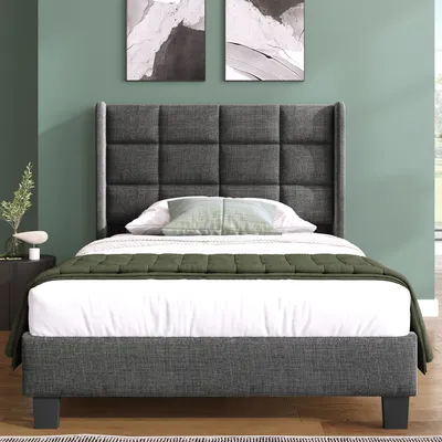 A bed with a grey upholstered headboard and frame, dressed with white bedding and a green quilted throw, set against a green wall with two abstract art pieces.
