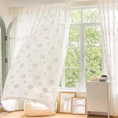 A bright room with a large window covered by sheer curtains with a cloud pattern. A small cabinet and framed pictures are visible.
