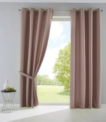 Two taupe curtains are shown hanging in front of a window with a view of a grassy field and trees. A small side table with a pineapple and two apples is to the left of the curtains.