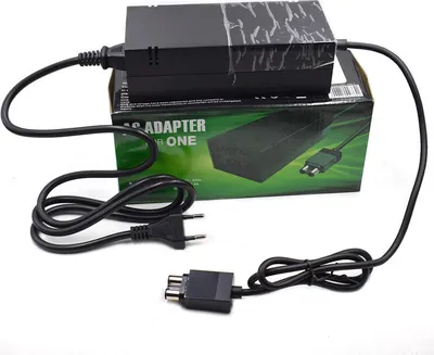 A black Xbox One power adapter and its cables are laid out on a white surface, with the product's packaging box in the background.