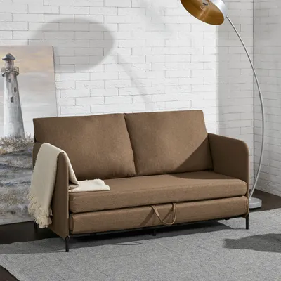 A brown sofa with a throw blanket draped over the armrest, placed in a room with a white brick wall and a floor lamp.