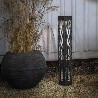 A black solar-powered pathway light with a decorative, branch-like pattern stands on a gravel surface next to a large, dark, textured planter filled with dried grass.
