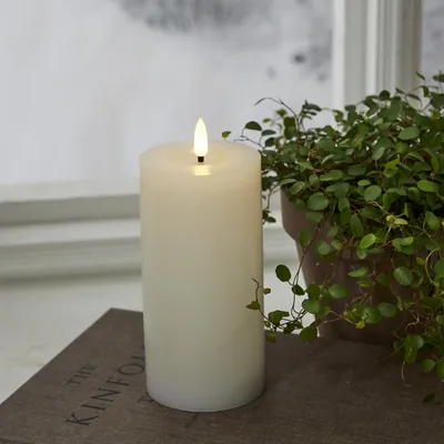 A white LED candle with a flickering flame sits on a book next to a potted plant, with a blurred window in the background.