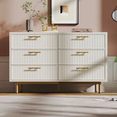 A white dresser with gold legs and handles is shown in a room with pink walls and a rug.