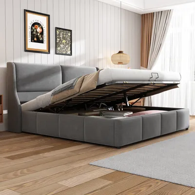 A grey upholstered bed with its storage compartment open, revealing items inside. The bed is in a room with wooden flooring and artwork on the wall.