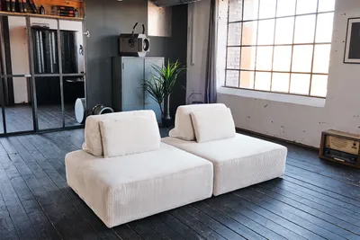 Two cream-colored modular sofa elements are placed on a dark wooden floor in a loft-style room with large windows.