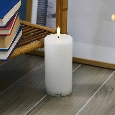 A white LED candle with a flame sits on a wooden floor next to a stack of books and a bamboo shelf.