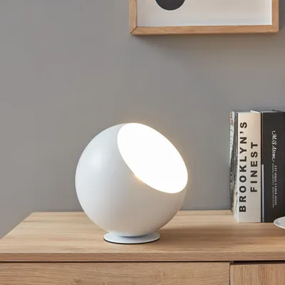 A white spherical table lamp with a cut-out section emitting light, placed on a wooden table next to two books.