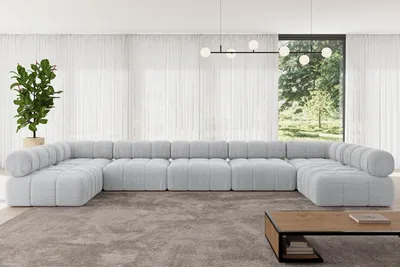 A large, light gray modular U-shaped sofa is shown in a modern living room with sheer white curtains and a large window overlooking trees. A coffee table is in the foreground.