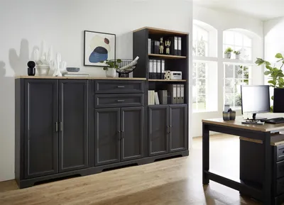 A modern office setting with a large dark gray cabinet and bookshelf unit, a wooden desk with a computer, and large windows in the background.