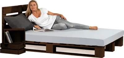 A woman is lying on a bed frame made of wooden pallets, reading a book.