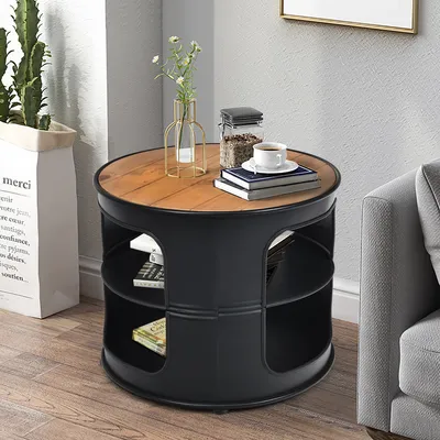 A round side table with a wooden top and black metal frame, featuring shelves, is placed next to a sofa and a large potted plant. A cup of coffee and books are on the table.