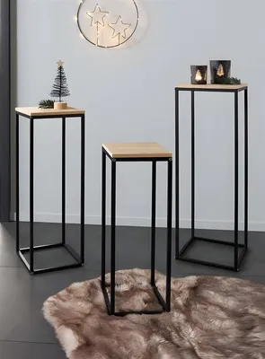 Three modern side tables of varying heights are arranged on a fluffy rug in front of a white wall. The tables have wooden tops and black metal frames. The tallest table on the right has two lit candles and a sprig of greenery. The middle table is shorter a