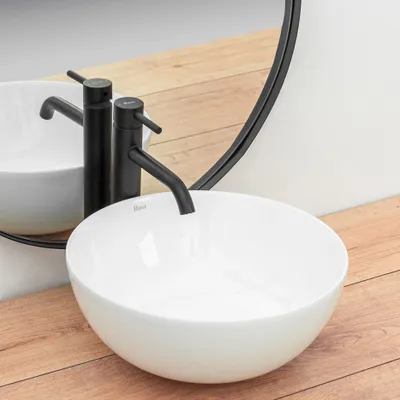 A white ceramic basin with a black faucet is shown on a wooden countertop, with a large round mirror in the background.